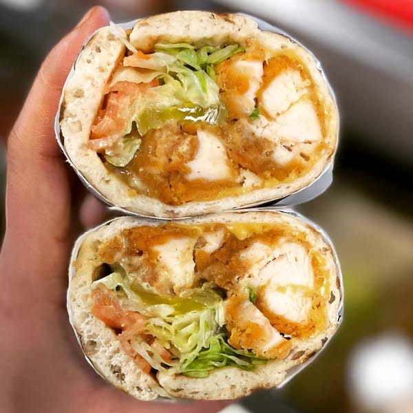 A hand holds a sliced chicken sandwich from Delite Cafe and Deli Taylor, revealing crispy chicken, lettuce, and tomatoes.