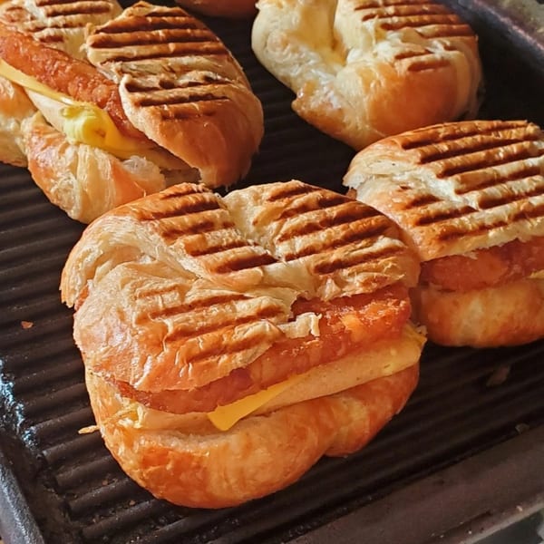 Several grilled croissant sandwiches with sausage and cheese on a panini press at Delite Cafe and Deli Taylor.